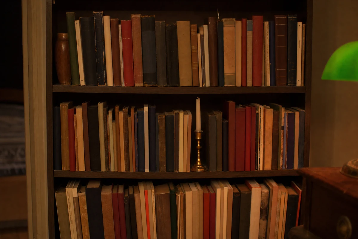 A dark-brown bookcase filled edge to edge with hardcovers, several with their paper jackets removed to show navy and dark red cloth, one brass candlestick holding an unlit taper on a shelf, and a green-shaded lamp lit beside the case