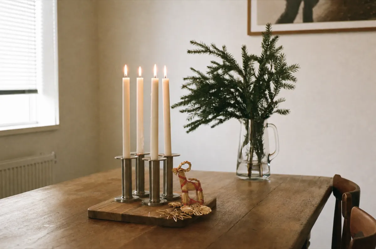 Four lit white taper candles in plain steel holders on a wood board, with straw ornaments and a straw goat beside them and a jug of fir branches well behind